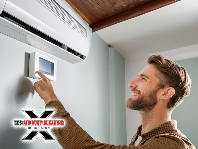 man standing adjusting thermostat on wall smiling inside residential home with ceiling clean air ducts visible behind him lower energy bills